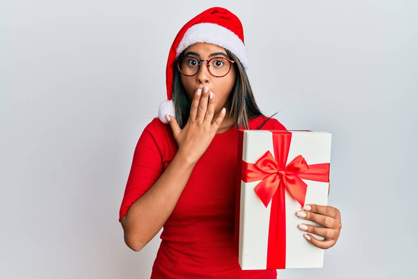 Молода латинська дівчина носить різдвяний капелюх і тримає подарунок, що покриває рот рукою, шокований і боїться помилки. здивований вираз
  - Фото, зображення