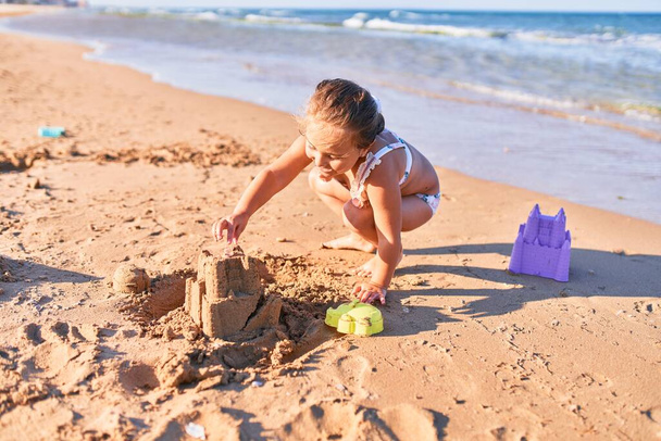 Urocze blond dziecko w bikini. Budowa zamku z piasku przy użyciu wiadra i łopaty na plaży - Zdjęcie, obraz