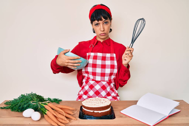 Mooie brunettte vrouw koken worteltaart met behulp van garde sceptisch en nerveus, fronsen boos vanwege probleem. negatieve persoon.  - Foto, afbeelding