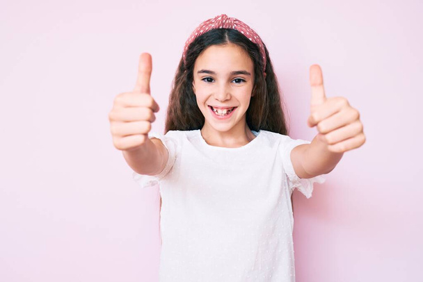 Cute hispanic child girl wearing casual clothes and diadem approving doing positive gesture with hand, thumbs up smiling and happy for success. winner gesture.  - Fotografie, Obrázek