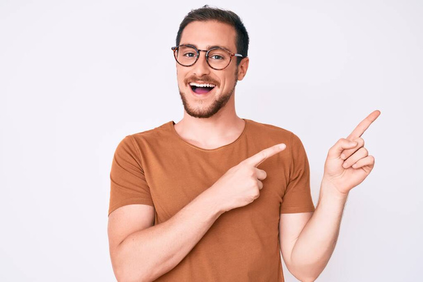 Νεαρός όμορφος άντρας που φοράει καθημερινά ρούχα και γυαλιά χαμογελώντας και κοιτάζοντας την κάμερα που δείχνει με τα δύο χέρια και τα δάχτυλα στο πλάι.  - Φωτογραφία, εικόνα
