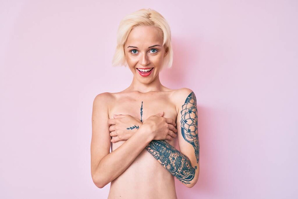 Young blonde woman with tattoo standing shirtless smiling and laughing hard out loud because funny crazy joke.  - Photo, image
