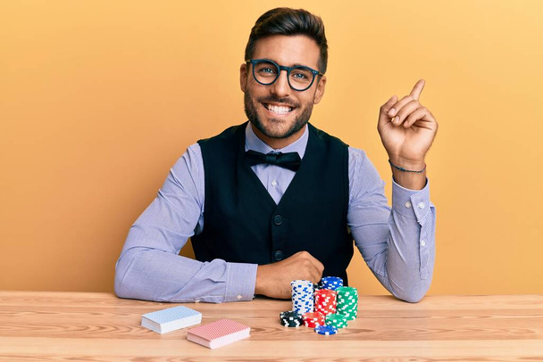 Hezký hispánský krupiér sedí na stole s pokerovými žetony a kartami a šťastně ukazuje rukou a prstem na stranu  - Fotografie, Obrázek