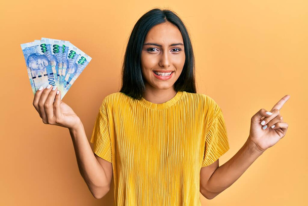 Young brunette woman holding south african 100 rand banknotes smiling happy pointing with hand and finger to the side  - 写真・画像
