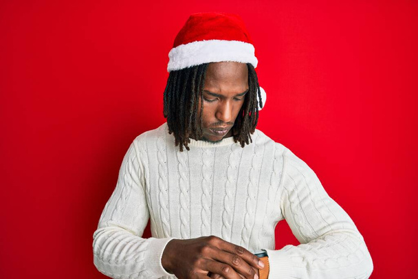 Afro-Amerikaanse man met vlechten dragen kerstmuts controleren van de tijd op de pols horloge, ontspannen en zelfverzekerd  - Foto, afbeelding