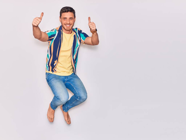 Jonge knappe Latijns-Amerikaanse man met casual kleding die vrolijk lacht. Springen met een glimlach op het gezicht doet ok teken met duimen omhoog over geïsoleerde witte achtergrond - Foto, afbeelding