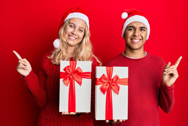 Νεαρό διαφυλετικό ζευγάρι φοράει χριστουγεννιάτικο καπέλο και κρατώντας ένα δώρο χαμογελώντας χαρούμενος δείχνοντας με το χέρι και το δάχτυλο στο πλάι  - Φωτογραφία, εικόνα