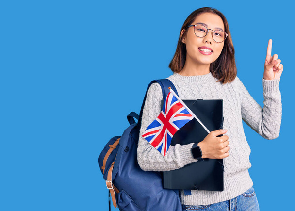 Fiatal gyönyörű kínai lány visel diák hátizsák kezében kötőanyag és UK zászló meglepett egy ötlet vagy kérdés ujjal mutogató boldog arc, első számú  - Fotó, kép