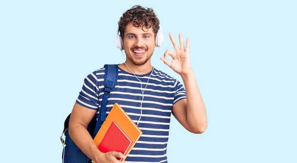 Joven hombre guapo con el pelo rizado sosteniendo mochila estudiante y libros haciendo signo bien con los dedos, sonriendo gesto amistoso excelente símbolo  - Foto, imagen