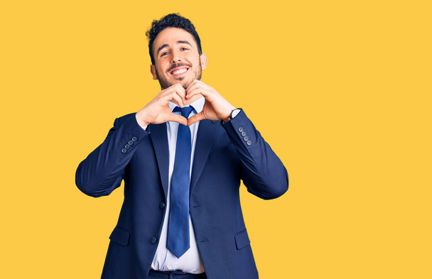 Junger hispanischer Mann in Businesskleidung, der verliebt lächelt und Herzsymbol und Form mit den Händen zeigt. Romantisches Konzept.  - Foto, Bild