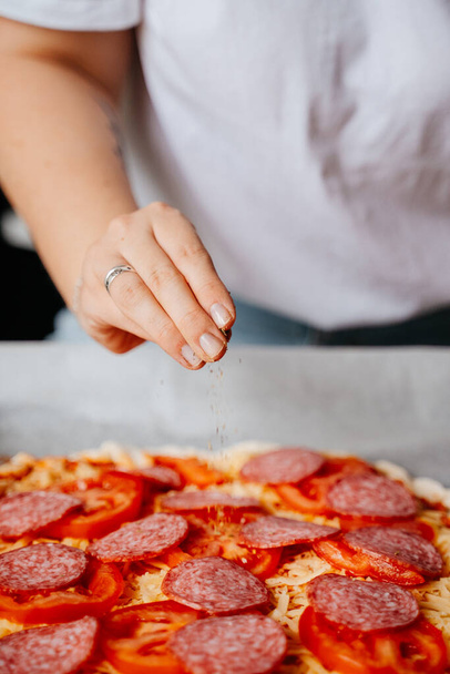 Gros plan d'une main versant du poivre et des épices aux herbes dans une pizza pepperoni. Préparation de la nourriture maison pour rencontrer des amis. - Photo, image