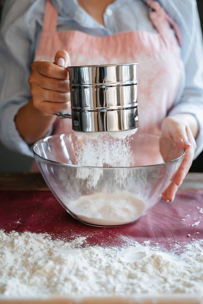 Młoda dziewczyna gotuje w fartuchu przesiewa białą mąkę pszenną do szklanej miski. Przygotowanie ciasta do pieczenia khachapuri. Sito żelazne do mąki. - Zdjęcie, obraz
