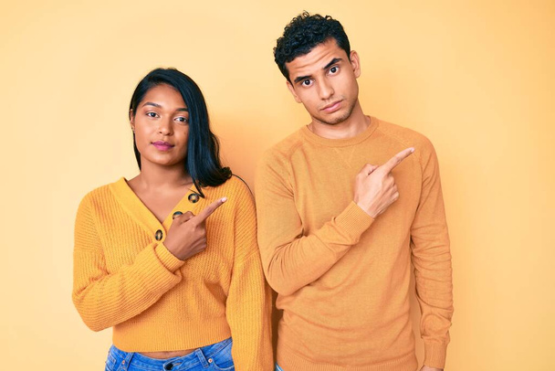 Beautiful latin young couple wearing casual clothes together pointing with hand finger to the side showing advertisement, serious and calm face  - Valokuva, kuva