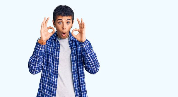 Young african amercian man wearing casual clothes looking surprised and shocked doing ok approval symbol with fingers. crazy expression  - Фото, зображення