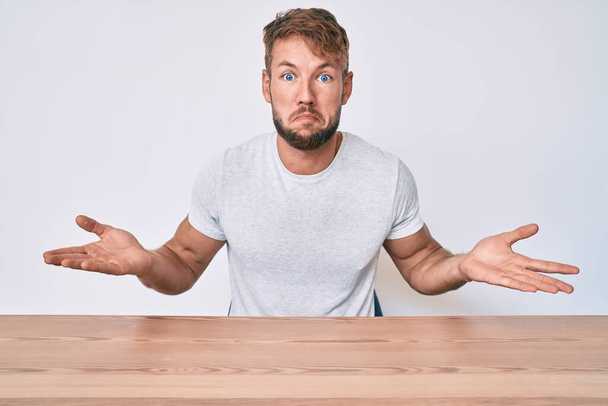 Young caucasian man wearing casual clothes sitting on the table clueless and confused expression with arms and hands raised. doubt concept.  - Photo, Image