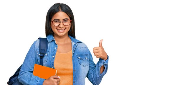 Menina latina usando mochila estudantil e segurando livros fazendo polegares felizes gesto com a mão. aprovando expressão olhando para a câmera mostrando sucesso.  - Foto, Imagem