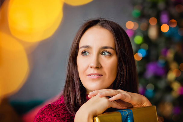 Portrait of a dark-haired girl with a gift on the background of a Christmas tree. - Foto, Imagem