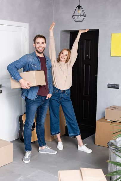 homme tenant boîte en carton près de femme heureuse debout avec les mains au-dessus de la tête, concept de relocalisation - Photo, image