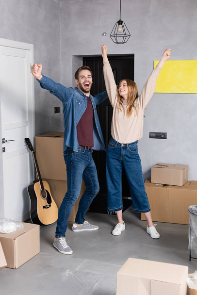 excited couple standing with hands above head near carton boxes, relocation concept - Fotó, kép