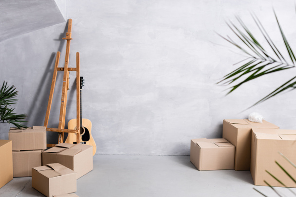 cajas de cartón cerca de caballete, guitarra acústica y plantas  - Foto, Imagen