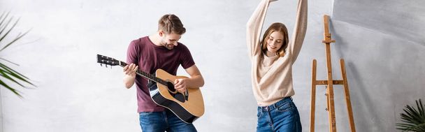 panoramatický záběr muže hrající na akustickou kytaru poblíž potěšené ženy tančící s rukama nad hlavou - Fotografie, Obrázek
