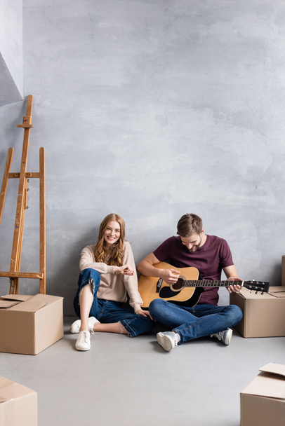 homem tocando guitarra acústica perto de mulher satisfeita sentado perto cavalete e caixas, conceito em movimento - Foto, Imagem