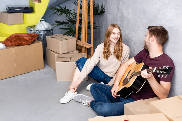 ευχαριστημένη γυναίκα κοιτάζοντας φίλο παίζει ακουστική κιθάρα κοντά σε κουτιά, έννοια μετεγκατάσταση - Φωτογραφία, εικόνα