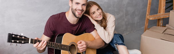 horizontaal beeld van tevreden vrouw knuffelen vriendje spelen akoestische gitaar in de buurt van dozen, verplaatsing concept - Foto, afbeelding