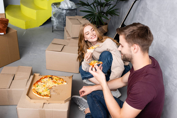 selective focus of joyful woman looking at man with pizza near carton boxes on floor - Photo, Image