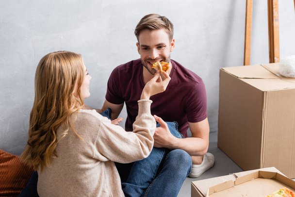young woman feeding bearded boyfriend with pizza near boxes - 写真・画像