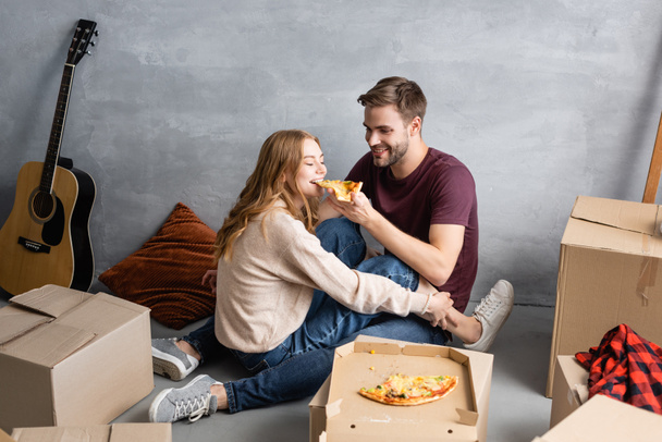 pleased man feeding girlfriend with pizza near carton boxes  - Fotoğraf, Görsel