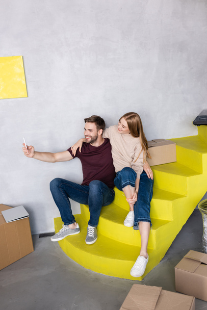 complacida pareja joven sentada en escaleras amarillas y tomando selfie cerca de cajas de cartón - Foto, imagen