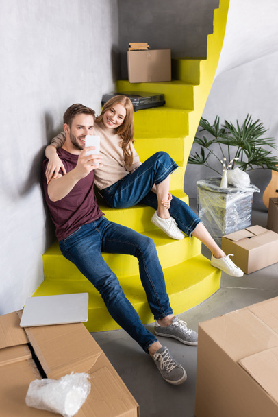pareja complacida sentada en escaleras amarillas y tomando selfie cerca de cajas de cartón - Foto, Imagen