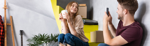 horizontales Bild eines fröhlichen Mannes, der Freundin mit Tasse fotografiert, während er auf Treppen in der Nähe von Schachteln sitzt - Foto, Bild