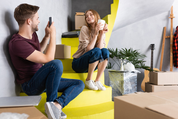 joyful man taking photo of girlfriend with cup while sitting on stairs near boxes - Photo, Image
