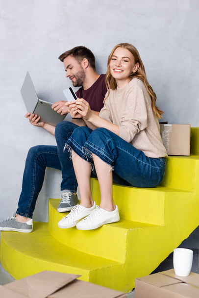 young woman holding credit card and sitting on stairs near man using laptop, relocation concept - Фото, изображение