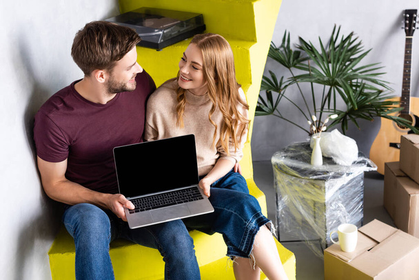 freudiges Paar hält Laptop mit leerem Bildschirm, während es auf Treppen in der Nähe von Boxen sitzt und einander anschaut, Umzugskonzept  - Foto, Bild