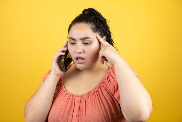 Молода красива жінка з кучерявим волоссям над ізольованим жовтим тлом розмовляє зі своїм телефоном, здивована, і у неї є ідея
 - Фото, зображення