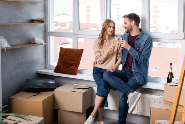 pleased man hugging girlfriend and clinking glasses with champagne near carton boxes, moving concept  - Photo, Image