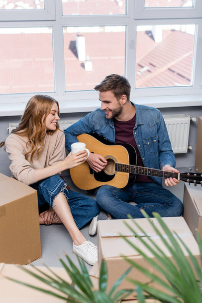 foyer sélectif de jeune homme jouant de la guitare acoustique près de la femme avec des boîtes de tasse et de carton  - Photo, image