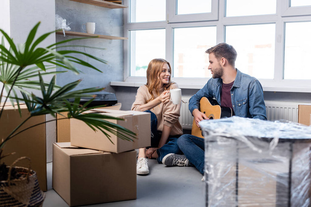 selective focus of young man playing acoustic guitar near pleased woman with cup and carton boxes  - Photo, Image