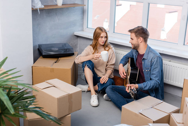 女性の近くでアコースティックギターを弾きながらカップとカートンの箱で座っている若い男の選択的な焦点  - 写真・画像