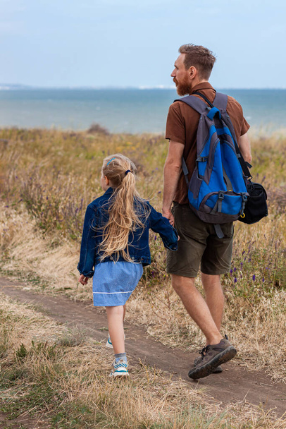 Батько з дочкою за руку стоїть на пагорбі з природним пейзажем. Маленька блондинка обіймається з татом на відкритому повітрі. Любляча дитина обіймає свого тата. Вітальна листівка до Дня батька
. - Фото, зображення