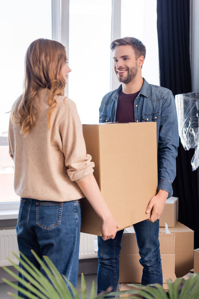 hombre alegre mirando a la mujer mientras sostiene caja de cartón juntos, concepto en movimiento - Foto, Imagen