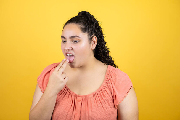 Young beautiful woman with curly hair over isolated yellow background disgusted with her hand inside her mouth - Photo, Image