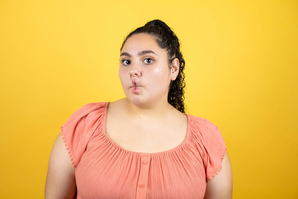 Jonge mooie vrouw met krullend haar over geïsoleerde gele achtergrond maken vis gezicht met lippen, gek en komisch gebaar. - Foto, afbeelding