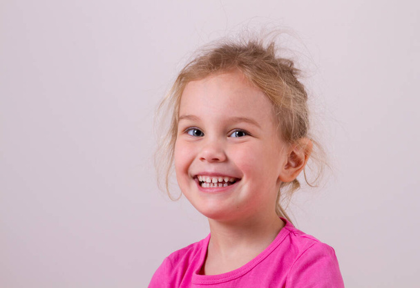 Close up shot of beautiful blonde Caucasian little girl wearing pink t-shit, looking at the camera with adorable smile, posing against white copy space concrete wall. Happy childhood concept - Φωτογραφία, εικόνα