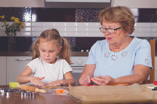 Grootmoeder met haar kleindochter bakt koekjes - gerold deeg op een tafel en gesneden koekjesvormen. Lachen. Veel plezier. - Foto, afbeelding