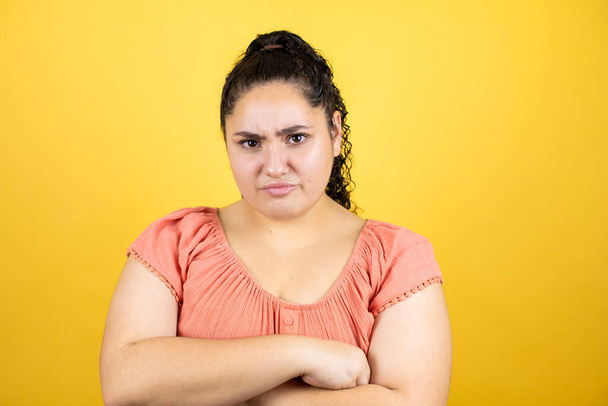 Jonge mooie vrouw met krullend haar over geïsoleerde gele achtergrond sceptisch en nerveus, afkeurende uitdrukking op gezicht met gekruiste armen - Foto, afbeelding