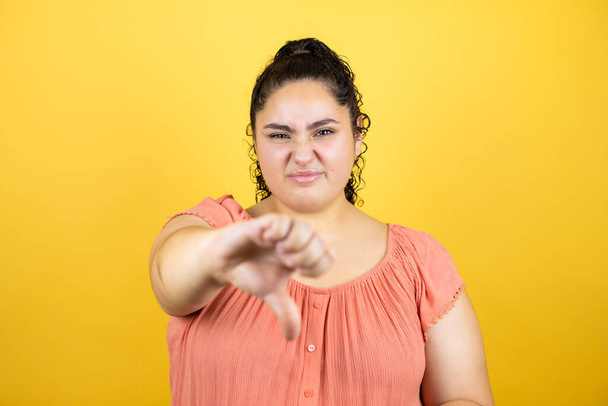Mladá krásná žena s kudrnatými vlasy přes izolované žluté pozadí s rozzlobeným obličejem, negativní znamení ukazující odpor s palcem dolů - Fotografie, Obrázek